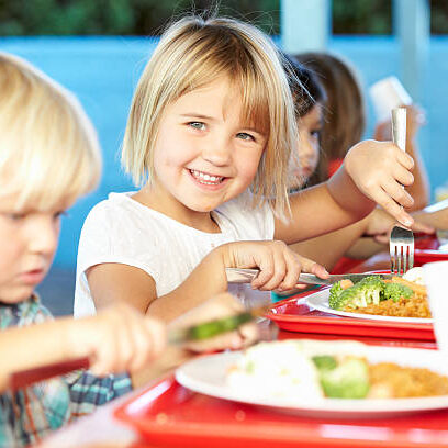 Elementary Pupils Enjoying Healthy Lunch In Cafeteria Sitting Down Smiling At Camera